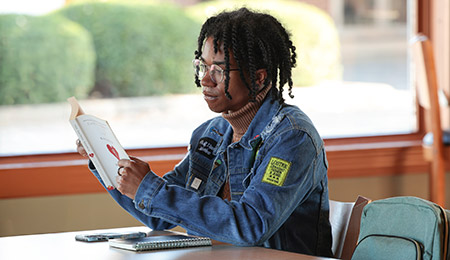 A student sits at a table by a large window reading a book, with a notebook, pen, and smartphone placed on the table. The student wears glasses, a denim jacket with patches, and headphones, and carries a backpack resting nearby.